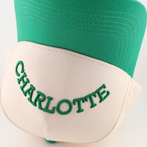 Op de afbeelding: Een witte en groene baseballpet met het woord "CHARLOTTE" in het groen geborduurd op de voorkant.