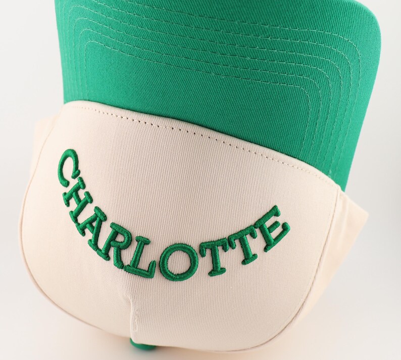 Op de afbeelding: Een witte en groene baseballpet met het woord "CHARLOTTE" in het groen geborduurd op de voorkant.