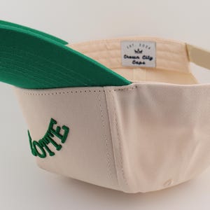 Op de afbeelding: Een cr&egrave;mekleurige baseballpet met een groene klep en een groen geborduurd woord "LOTTE" aan de zijkant. De pet heeft een wit label met de tekst "Crown City Caps" aan de achterkant.
