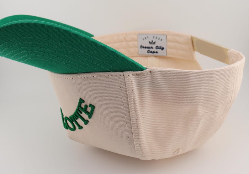 Op de afbeelding: Een cr&egrave;mekleurige baseballpet met een groene klep en een groen geborduurd woord "LOTTE" aan de zijkant. De pet heeft een wit label met de tekst "Crown City Caps" aan de achterkant.
