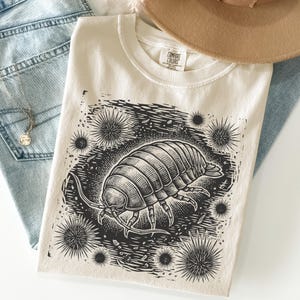 May include: A cream-coloured short-sleeve t-shirt featuring a black ink-style graphic of a pill bug surrounded by spiky sea urchin-like shapes. The shirt has a crew neckline and a Comfort Colors brand label printed on the inside of the collar.