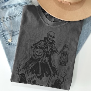May include: A dark grey t-shirt with a black illustration of a skeleton holding a jack-o'-lantern and a lantern. The skeleton wears a cape and stands before tombstones. A tan hat and blue jeans are in the background.