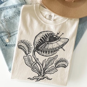 May include: Cream-colored t-shirt featuring a black graphic of a Venus flytrap with a UFO design. The shirt has a crew neck and short sleeves. The Comfort Colors brand tag is visible on the shirt.