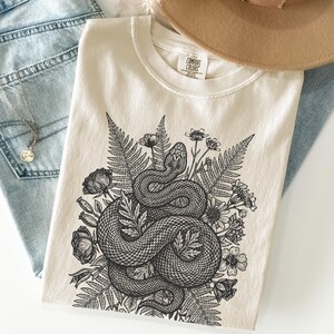 May include: Cream-colored t-shirt featuring a detailed black and white illustration of a coiled snake surrounded by ferns and flowers. The shirt has a crew neck and the brand name "COMFORT COLORS" is visible on the tag.