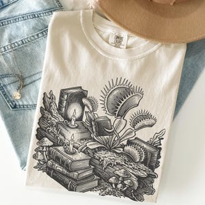 May include: Cream Comfort Colors t-shirt with a black and white illustration of books, candles, mushrooms, and a Venus flytrap. The shirt is folded on blue jeans and a brown hat. The design is detailed and artistic.