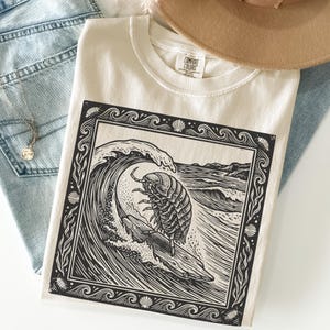 May include: Cream-colored t-shirt featuring a black and white graphic of a pill bug surfing a wave. The design is framed with a decorative border. The shirt is folded on top of blue jeans and a brown hat.