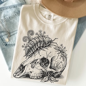 May include: A cream-colored t-shirt featuring a black ink graphic of an isopod crawling over an animal skull. The skull is adorned with small flowers and leafy vines. The shirt has a Comfort Colors brand label printed on the inside of the neck.