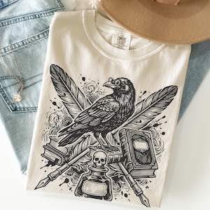 May include: Cream-colored t-shirt featuring a detailed black illustration of a raven wearing spectacles, crossed quill pens, feathers, books, and a skull. The shirt has the Comfort Colors brand tag.