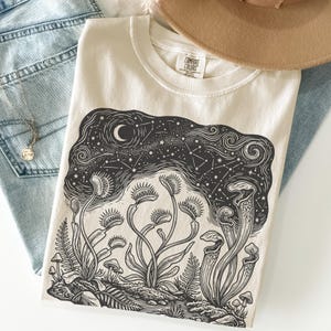 May include: Cream-coloured t-shirt with a black and white illustration of Venus flytraps, ferns, and mushrooms under a starry night sky and a crescent moon. The shirt is made of a soft material and has a crew neck.