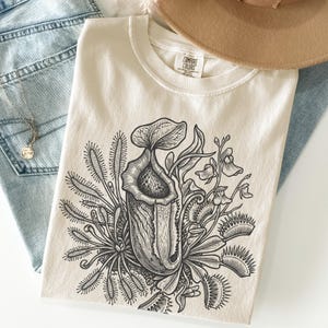 May include: Cream-colored t-shirt featuring a detailed black and white illustration of carnivorous plants, including a pitcher plant and Venus flytraps. The shirt has a crew neck and the Comfort Colors brand tag is visible.