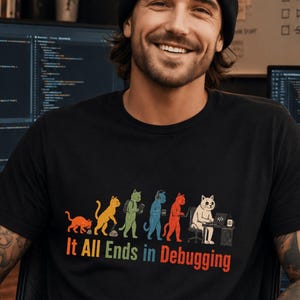 May include: A black short-sleeved t-shirt featuring a graphic of cats evolving into a programmer sitting at a computer. The text below the cats reads, It All Ends in Debugging, with each word in a different color including orange, yellow, green, and red.