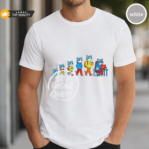 May include: A white short-sleeved t-shirt featuring a graphic of six blue cats in a developmental sequence, starting from a small kitten and progressing to a cat sitting at a desk with a laptop. The text 100% ORIGINAL QUALITY is printed below.