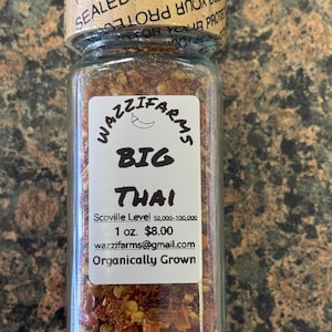 May include: A clear glass jar filled with red pepper flakes. The label reads "WAZZIFARMS BIG THAI" with the Scoville level and "Organically Grown" text. The jar is sealed with a lid that says "SEALED FOR YOUR PROTECTION".