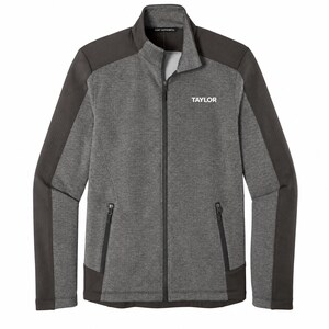 Puede incluir: Una chaqueta gris carbón con cremallera completa, cuello alto, mangas largas con paneles gris oscuro y dos bolsillos laterales con cremallera. La palabra TAYLOR está impresa en blanco en el pecho izquierdo y la marca Port Authority es visible en el cuello.