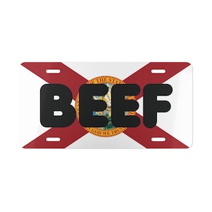 May include: A rectangular license plate featuring the Florida state flag design. The flag has a white background with a red X and the state seal in the center. Bold black letters spell out the word "BEEF" across the center of the plate.