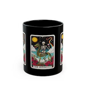 May include: Black ceramic mug featuring a tarot card design. The card depicts a skeleton riding a dragon, holding a glass of wine, with a rainbow and sun in the background. The card is surrounded by red roses, with the text "THE AUDACITY".