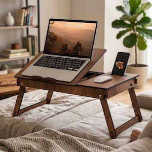 May include: A dark brown wooden laptop desk with foldable legs, featuring an adjustable angled platform for a laptop, a flat side section with a white computer mouse, and a dedicated slot holding a black smartphone with a floral screen display.