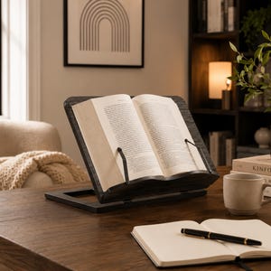 Puede incluir: Un atril de madera gris oscuro que sostiene un libro de tapa dura abierto con páginas blancas. El soporte cuenta con dos finos sujetapáginas de metal negro unidos al borde frontal para mantener el libro abierto. La base es un marco de madera plano.