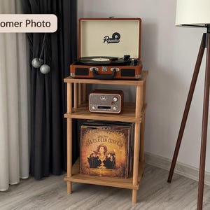 May include: A three-tiered light wood storage stand with four vertical legs. The top shelf holds a brown portable record player with the text Piranha. The middle shelf holds a small brown radio. The bottom shelf contains several vinyl records.