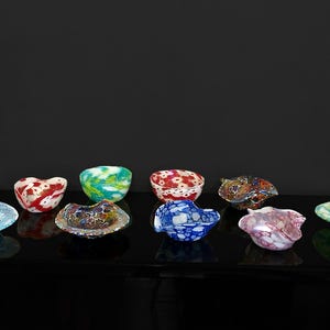 Può includere: Una collezione di ciotole decorative in vetro colorato. Le ciotole sono di varie forme e dimensioni, con una varietà di colori e motivi, tra cui rosso, blu, verde e viola. Le ciotole sono disposte su una superficie nera riflettente.