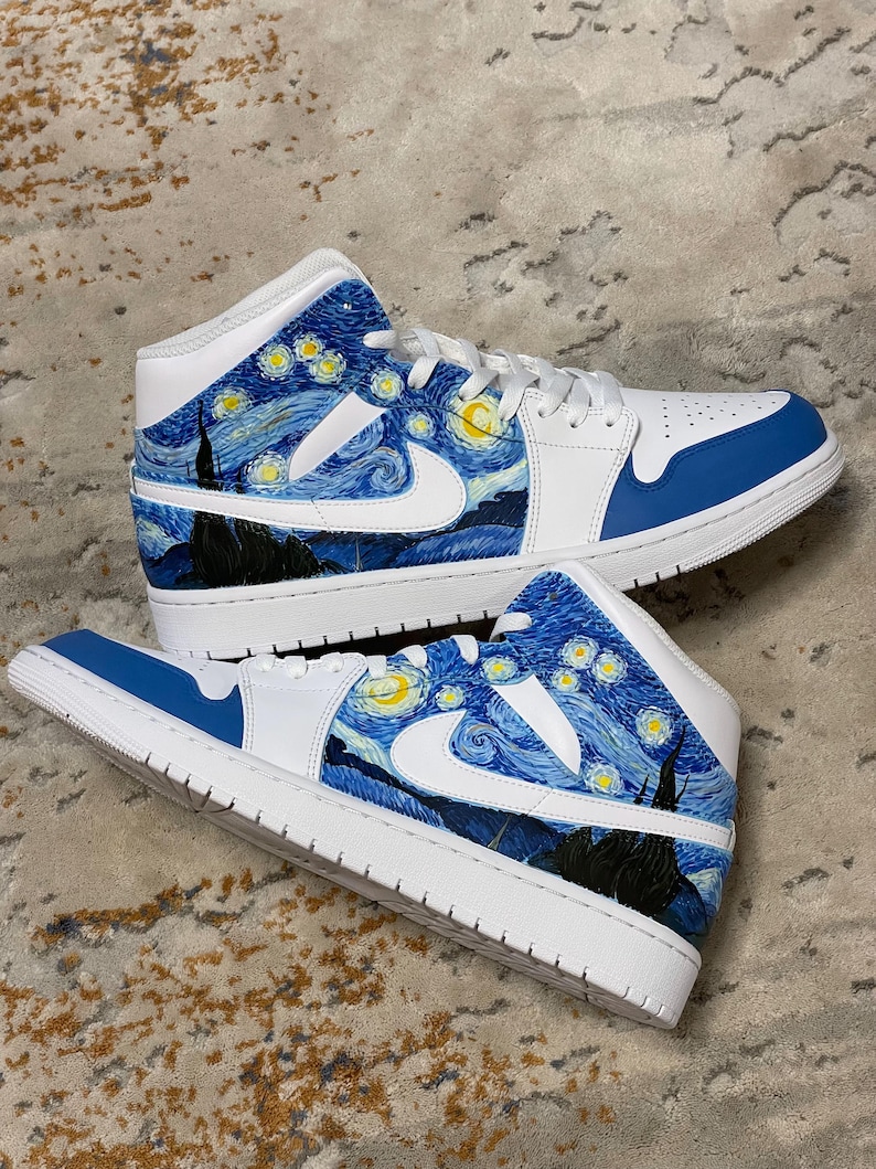 May include: Pair of white and blue sneakers with a custom design inspired by Van Gogh's "Starry Night" painting. The design features swirling blue brushstrokes, yellow stars, and a black silhouette of a village. The shoes have white laces and soles.