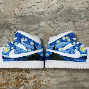May include: Pair of white and blue high-top sneakers featuring a starry night design. The sneakers have a white base with blue painted accents depicting a night sky with swirling clouds and stars. The Nike swoosh is white.