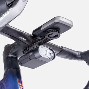 May include: A black Garmin cycling computer mounted on a carbon fiber handlebar stem, positioned above a black bicycle headlight with a silver circular lens housing. The stem features the BMC logo in white text on its side, and the setup is attached.