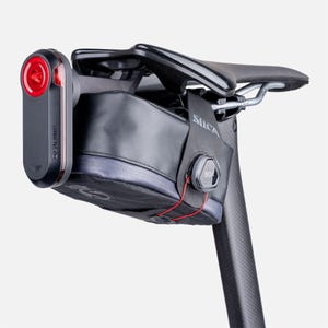 Könnte beinhalten: Ein schwarzer Fahrradsattel mit einer schwarzen Aufbewahrungstasche und einem roten Garmin-Licht. Die Tasche wird mit einem BOA-Drehsystem befestigt. Der Sattel ist auf einer Sattelstütze aus Kohlefaser montiert.