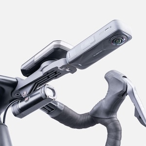 Op de afbeelding: Een fietsstuur met een gemonteerde camera, fietscomputer en koplamp. De camera is grijs en zwart, met een lens. De fietscomputer is zilver en zwart. De koplamp is zwart en cilindrisch. Het stuur is zwart met grijze tape.