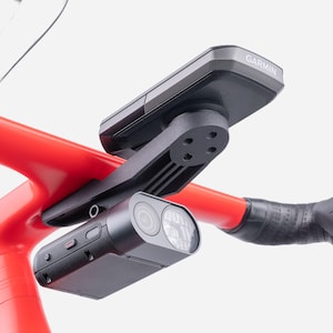 May include: A black Garmin cycling computer mounted on a black plastic bracket attached to a bright red bicycle handlebar. A black dual-lens camera is secured underneath the same mount, featuring a textured side panel with visible control buttons.