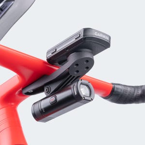 May include: A black Garmin cycling computer and a black cylindrical light are mounted on a red bicycle handlebar. The Garmin device is labelled with the brand name. The light has a lens and a red power button.
