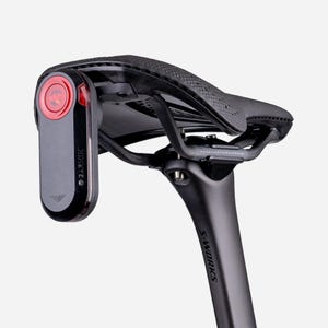 May include: A black bicycle saddle mounted on a matte black S-Works seatpost, featuring a vertical Garmin radar tail light attached to the rear. The light has a circular red lens at the top and a black casing with the Garmin logo printed on the side.