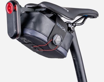 May include: A black bicycle saddle bag with a BOA closure system and the Silca logo, mounted under a black bike seat. A red and black Garmin rear light is attached to the back of the bag, which features a grey zipper and thin red cord.