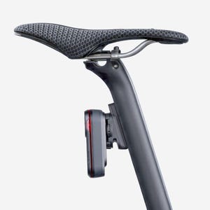 May include: A bicycle seat with a black and gray patterned design. A black seat post supports the seat, and a gray device with a red light is attached to the post. The device is a bicycle light.