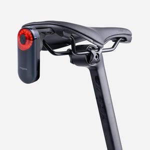 Puede incluir: Un sillín de bicicleta negro montado en una tija con una luz trasera LED roja de la marca Magene fijada en la parte posterior. La luz presenta una lente roja circular con un botón de encendido central y una carcasa gris oscuro bajo el asiento.
