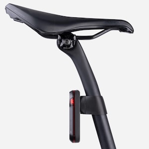 以下が含まれることがあります： 上面に「POWER」の文字がプリントされた黒い自転車のサドル。黒いシートポストがサドルに取り付けられています。赤いライトが付いた黒いGarminテールライトがシートポストに取り付けられています。