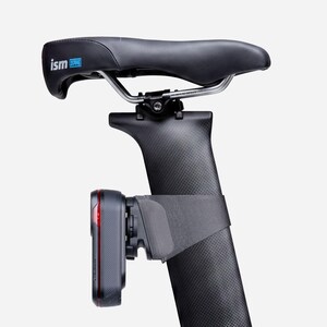 May include: A black bicycle seat with the brand "ism" printed on it. The seat is mounted on a black carbon fiber seat post. A black and gray device with red accents is attached to the seat post.