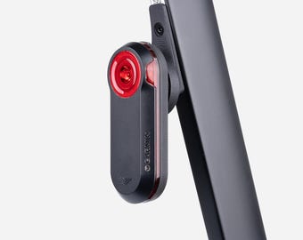 May include: A black Garmin bicycle tail light with a circular red lens and a vertical red light strip, mounted onto a black seat post. The device features a matte black casing with the Garmin brand name printed in small white letters on the side.