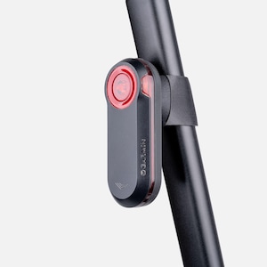 Può includere: Una luce posteriore radar per bicicletta Garmin nera con una lente rossa circolare sulla parte superiore e una striscia luminosa rossa verticale sul lato. Il dispositivo è montato su un reggisella cilindrico nero tramite una cinghia in gomma.