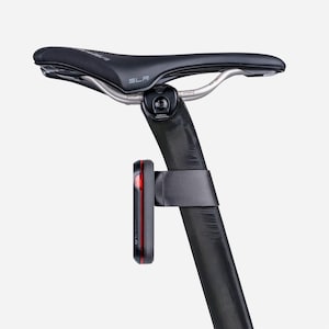 May include: A black bicycle seat with silver rails and a black seat post. A red and black bicycle tail light is attached to the seat post with a black strap. The seat has the word "SLR" printed on it.