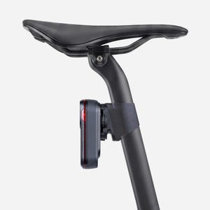 May include: A black bicycle saddle mounted on a seat post with a rectangular black and red rear safety light attached to the post. The light features a textured dark gray casing with a thin red vertical strip visible on the side of the unit.