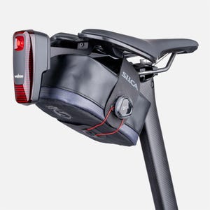May include: A black bicycle saddle bag with a BOA dial closure system and the Silca logo, mounted under a bicycle seat. A Wahoo brand red rear light is attached to the back of the bag, featuring a vertical red lens and a black housing.