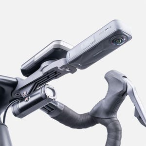 Pode incluir: Um guiador de bicicleta com uma câmara montada, um computador de ciclismo e um farol. A câmara é cinzenta e preta, com uma lente. O computador de ciclismo é prateado e preto. O farol é preto e cilíndrico. O guiador é preto com fita cinzenta.