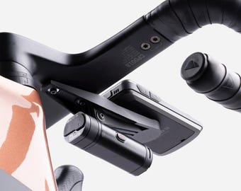 Può includere: Primo piano di un manubrio da bicicletta nero con accessori integrati. Una luce cilindrica nera e un dispositivo rettangolare sono montati sul manubrio. Il manubrio è avvolto con nastro nero. Il telaio della bici è visibile.