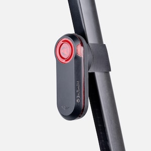 Op de afbeelding: Een zwart Garmin-fietsachterlicht met een ronde rode lens en een verticale rode lichtstrip aan de zijkant. Het apparaat is bevestigd aan een zwarte zadelpen met een zwarte rubberen band en is voorzien van de merknaam Garmin op de zijkant.