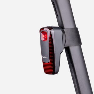 May include: A black Wahoo bicycle tail light with a red lens featuring horizontal stripes, mounted to a black seat post. The device has a rectangular shape with rounded edges and the Wahoo brand name printed in white on the front surface.