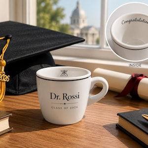 May include: A white ceramic mug with the text "Dr. Rossi CLASS OF 2026" and a black rim. A graduation cap with a gold tassel and the year 2026 is behind the mug. A rolled diploma and a small book are also visible.