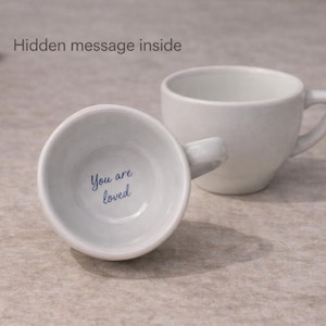 Può includere: Due tazze da caffè in ceramica bianca, una in posizione verticale e una inclinata, che rivelano il messaggio "You are loved" all'interno. Le tazze hanno un design semplice e moderno con manico. Il testo "Hidden message inside" è visibile.