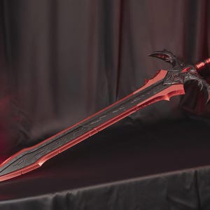 May include: A large sword featuring a black central blade with intricate engraved patterns, framed by sharp red edges. The hilt is black with ornate, wing-like crossguards and a red-accented grip, culminating in a pointed pommel at the end.