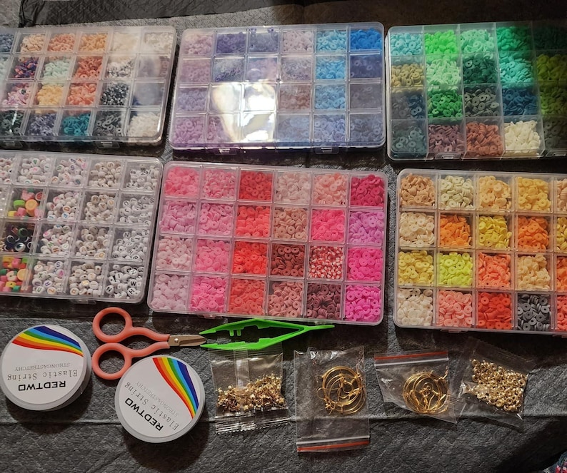 May include: A collection of jewelry-making supplies, including various colored beads in clear plastic organizers, spools of elastic string, scissors, tweezers, and small bags of gold-colored findings and hoop earrings. The beads are arranged by color, offering a wide range of options.